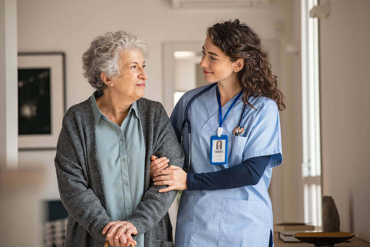 Young woman in light blue scrubs holding the arm of a senior woman in a gray cardigan.
