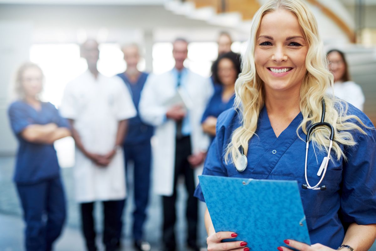 A nurse stands in the foreground, with healthcare colleagues in the background.