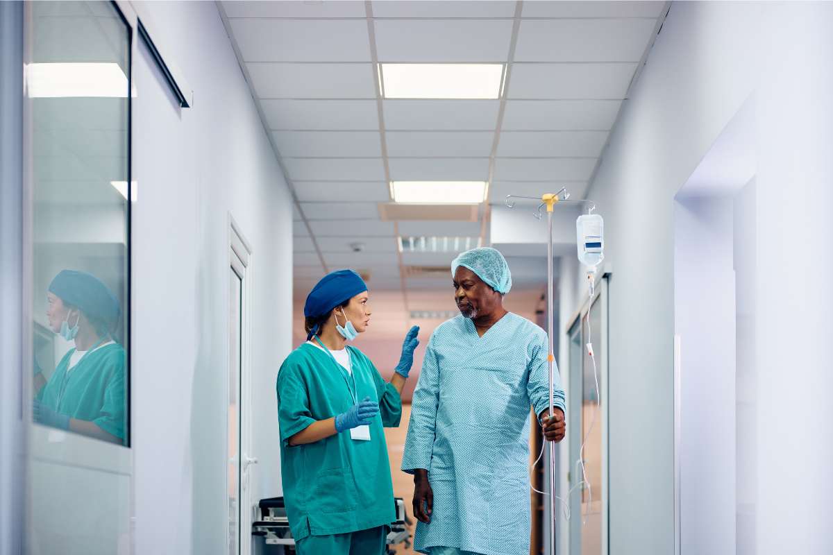 A nurse in scrubs walking down the hall of a hospital with a patient in a hospital gown.