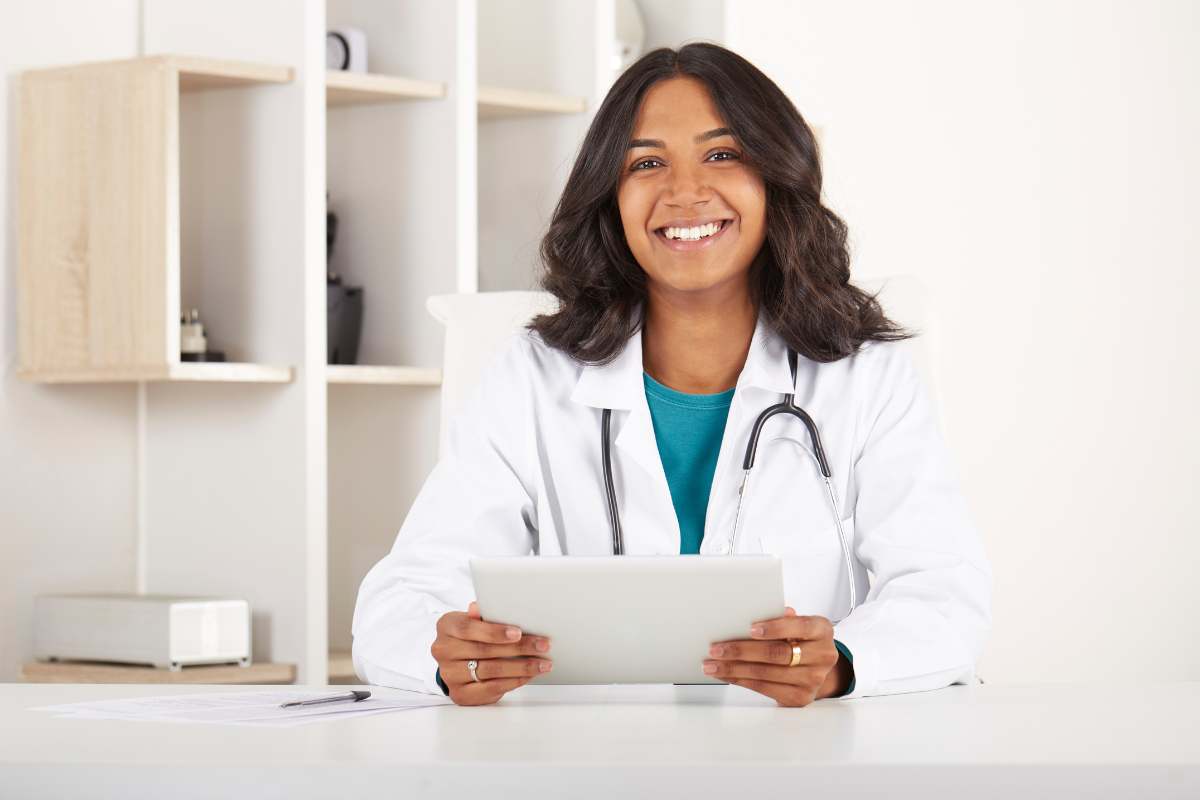 A female nurse practitioner in a white coat sitting at a desk, holding papers and smiling.