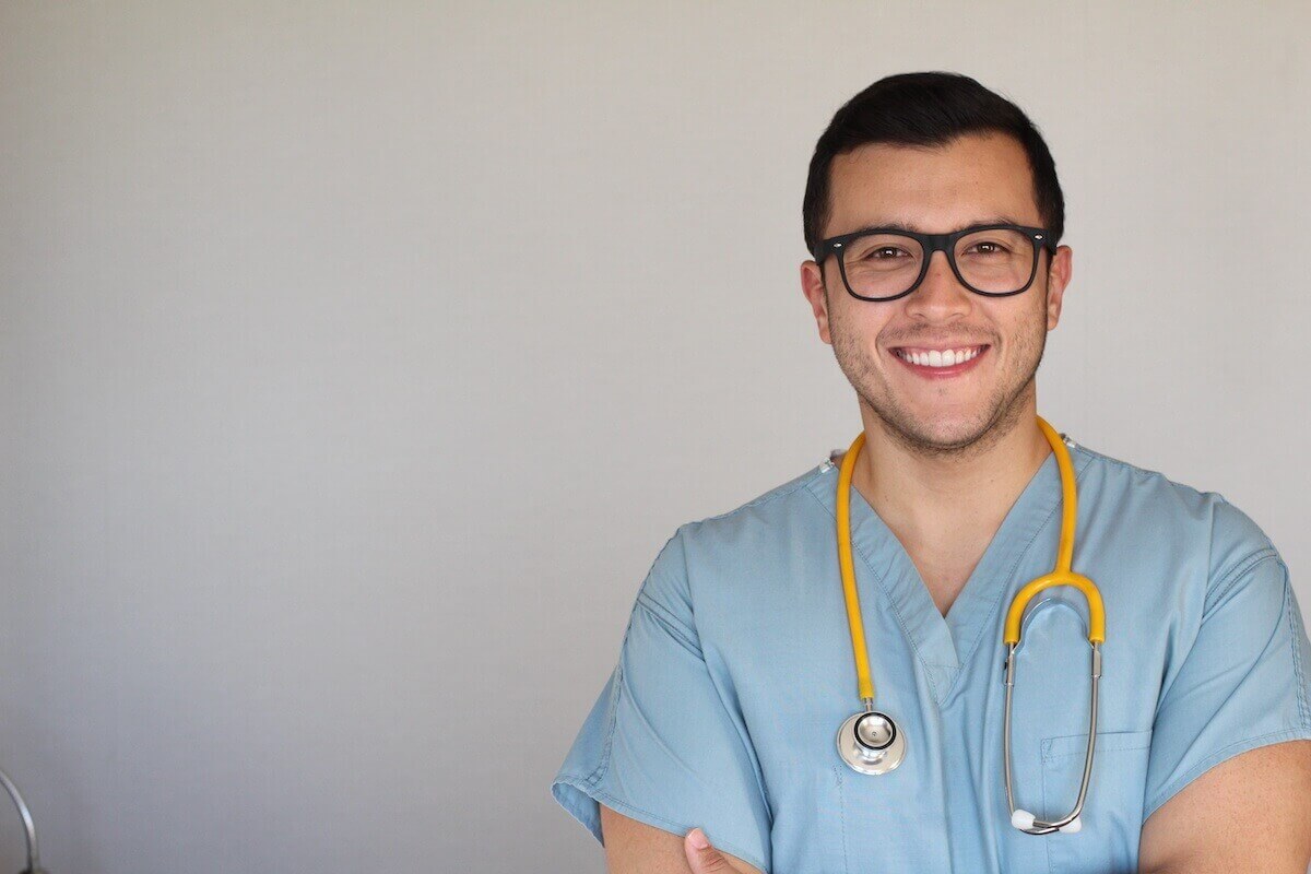 Young male nurse with dark hair and glasses in light blue scrubs.