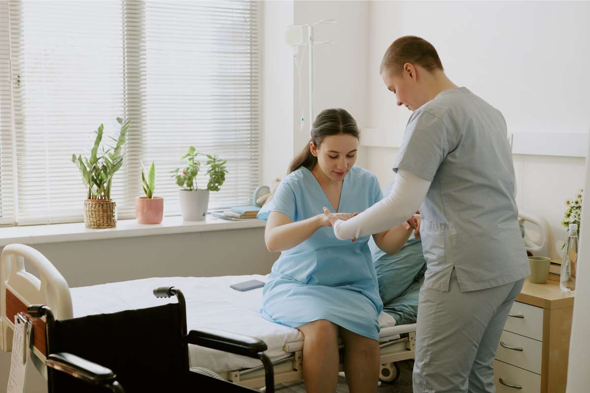 A nurse in gray scrubs helping a young female patient up from a hospital bed as part of nursing prioritization tasks.