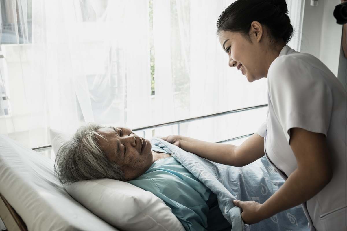 Elderly woman in hospital bed and young nurse putting a blanket on her to make her more comfortable.