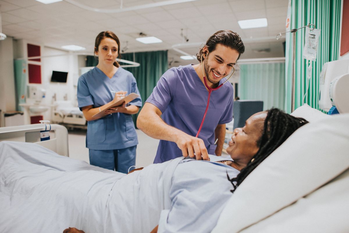 A nurse assists a patient while a CNA stands by and takes notes.