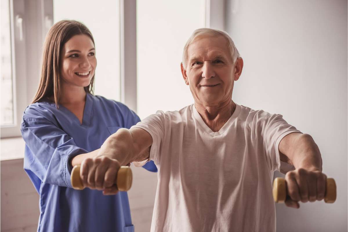 Female nurse in scrubs in a skilled nursing facility helping a patient properly lift weights as part of rehabilitation.